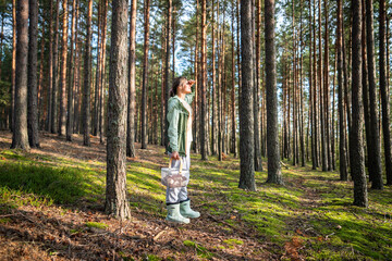 Fototapeta premium Relaxed woman with mushroom basket enjoying forest view looking forward, covering eyes in dense fall pine woodland. Female traveler with basket walk in woodland alone, mindfulness and slow living