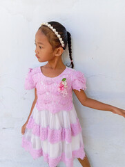 A little girl standing sideways in a lace dress, looking curious and natural against a simple background.