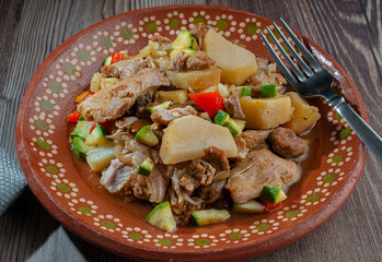 Mexican handmade clay dish with chicken stew and tomato sauce with potatoes, carrots, zucchini, and onions, with a fork on the side on a rustic wooden table