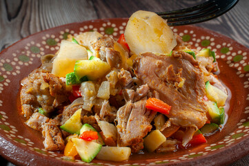 Mexican handmade clay dish with chicken stew and tomato sauce with potatoes, carrots, zucchini, and onions, with a fork picking up a potato, a rustic wooden table