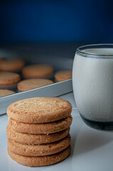 Stack of cookies next to a glass of cold milk and a tray of freshly baked cookies with a blue background. Foods high in sugars, saturated fat, and carbohydrates. Vertical.