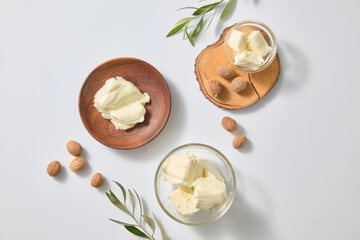 Simple overhead layout with glass jar of shea butter, whole nuts, wooden dish and olive twig against white background for natural skincare ingredient themes.