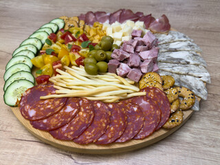 Charcuterie board with cucumber slices, chorizo, smoked ham, canned ham, head cheese, tomatoes, olives, cheese cubes, breadsticks and crackers on wooden table.