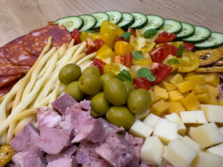 Close-up of charcuterie platter with cucumber slices, chorizo, cheese cubes, ham, green olives, tomatoes, breadsticks and crackers on wooden table.