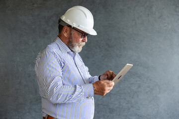 An older engineer wearing a white safety helmet uses a digital tablet, symbolizing modern construction practices, technology integration, and innovation in engineering.