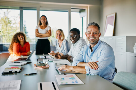 Diverse Business Team in a Bright Office Meeting Room. All looking into the camera smiling. They are sitting in a new, stylish office, with notebooks, paper and a laptop on the table