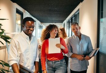 Diverse Team Enjoying A Casual Meeting In Modern Office Hallway. The colleagues walk together down the hallway, talking and laughing together. They are on their way to a meeting.