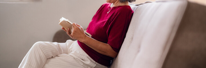 Older Asian woman reading book for relaxation at home living room on sofa cozy peaceful moment...