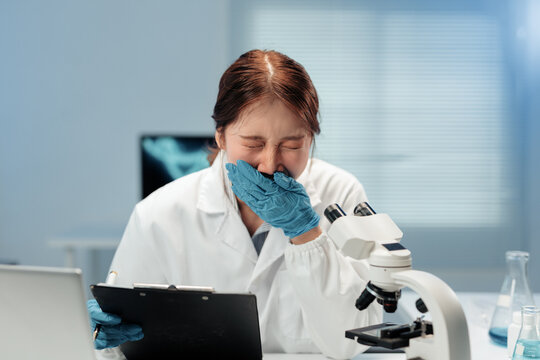 Young asian scientist woman wearing lab coat and gloves is crying and yawning in laboratory, holding clipboard and pen