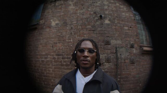 Young African American man wearing stylish 90s outfit with sunglasses speaking or rapping, old brick building in background, vignetting effect