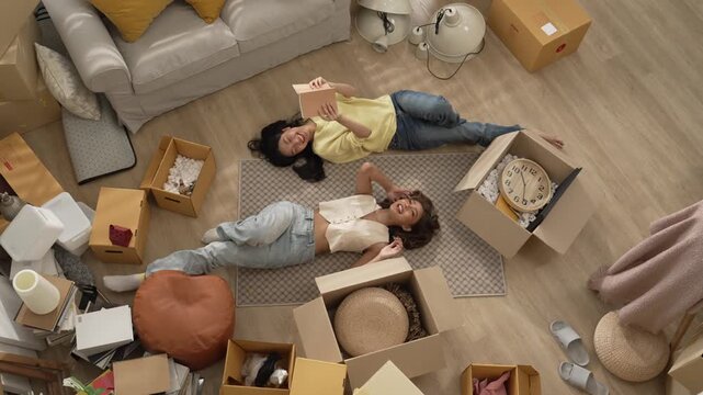 Smiling lesbian couple lying on the floor while spending time together. Top down view of girl lover reading a book while lying at living room surrounded with cardboard box in moving day. Myrmidon. - Powered by Adobe