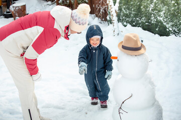 Grandmother and toddler building a snowman in snowy backyard. Joyful winter day, family bonding, healthy outdoor activity, seasonal lifestyle