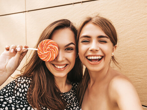 Fototapeta Two young smiling hipster women in summer clothes. Female taking selfie self portrait photos on smartphone.Models posing on street background near yellow wall.Eating candy lollipop