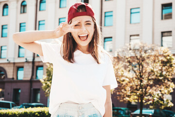 Young beautiful smiling hipster woman in trendy summer white t-shirt. Sexy carefree woman posing on the street background in cap at sunset.Positive model outdoors. Cheerful and happy.Shows peace sign
