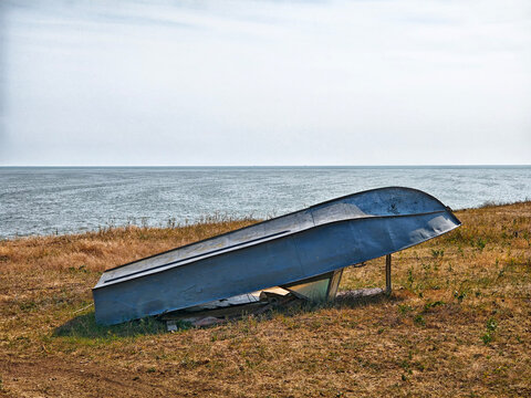Abandoned metal boat resting on a grassy shore beside calm waters under a clear sky at midday