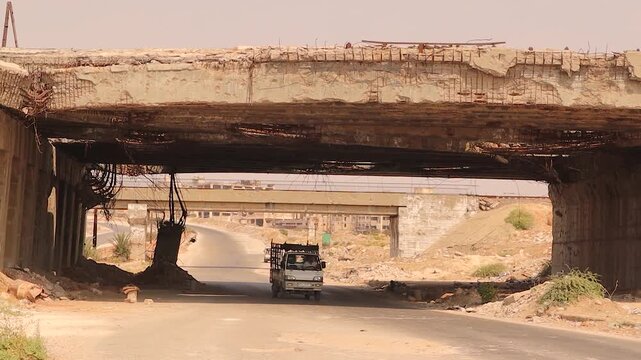 Destroyed and decayed bridge in Aleppo, Syria, with exposed steel bars and heavy war damage. A car passes beneath as if normal, reflecting how civilians adapt to war, resilience, courage and survival.
