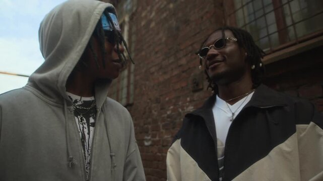 Low angle view of two cheerful African American guys wearing y2k streetstyle outfits chatting with friends near old brick building outdoors