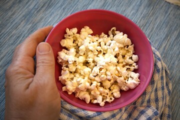 Close-up of popcorn in a pastel pink bowl placed on a blue checkered kitchen towel. Minimal composition with copy space and hand.