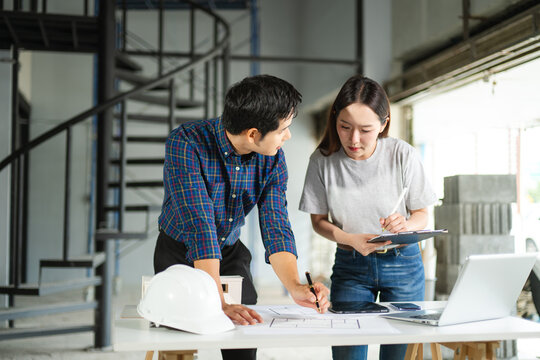 Asian male and female architects review blueprints at an indoor construction site. Teamwork, planning, and engineering in progress