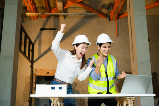 Two construction professionals in hard hats and safety vests raise their fists in celebration over completed project blueprints