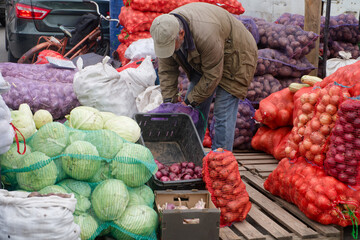 Unrecognizable vendor bends over bags of onions and cabbage at a street farmers market; rustic fall harvest trade scene with pallets and crates, candid reportage. Photo