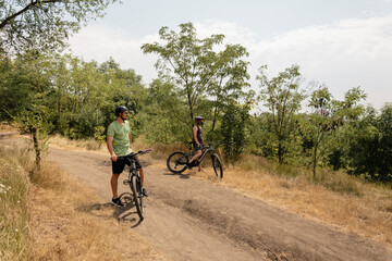 Two cyclists pause on a scenic trail surrounded by trees on a warm sunny day.