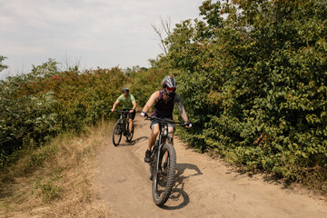 Two enthusiastic bikers riding on a dirt trail surrounded by lush green trees