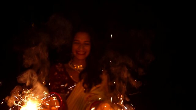Happy Indian woman celebrating Diwali festival with holding sparklers in hand. Lady wearing a traditional saree. Enjoying with fire crackers in dark night background.