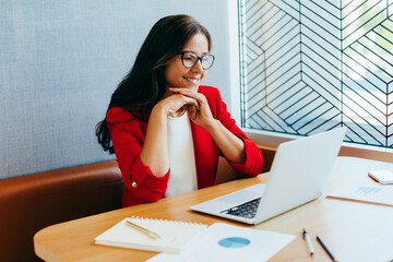 Smiling professional woman in red jacket working on laptop at office desk