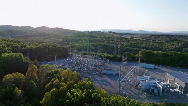 Electrical substation with high voltage transmission lines under evening sun in rural American landscape. Drone approaching shot. Green forest landscape in late summer.