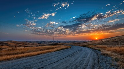 A curving gravel road winds through a dry, hilly landscape under a vibrant sunset sky, displaying a mix of blue and orange hues with scattered clouds