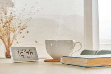 A cozy morning scene featuring a steaming cup of coffee next to a book and a digital clock, with soft natural light