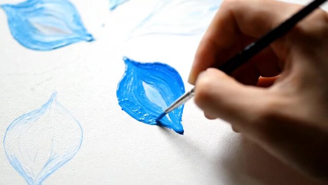 Close-up of an artist's hand painting a vibrant blue leaf with a fine brush on white paper