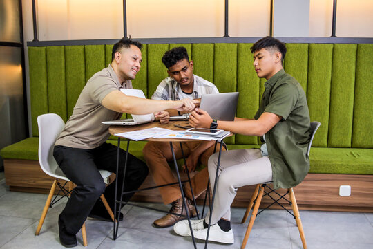 Three Young Men Collaborating on Project, Sitting at Table and Pointing at Laptop Screen