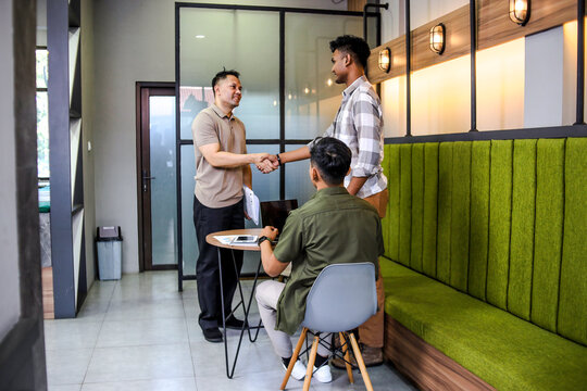 Male Colleagues Shake Hands in Modern Office While Third Colleague Sits at Table