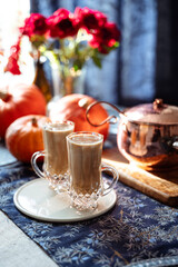 Two glass mugs of creamy pumpkin spice latte topped with cinnamon on a white plate. Autumn coffee drink served on a cozy festive table decorated with pumpkins, copper pot, and rustic details. 
