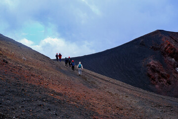 Group of hikers exploring volcanic slopes with dramatic red and black ash textures under cloudy sky, adventure trekking on active mountain terrain, perfect for travel, geology, and outdoor exploration