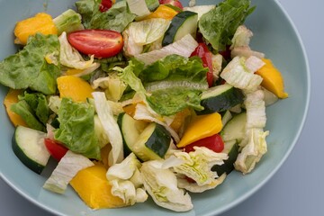 Bowl of mixed salad with lettuce leaves, cucumbers, cherry tomatoes, and mango. Healthy vegetarian food rich in vitamins, fresh organic meal for diet and wellness, isolated on gray background.