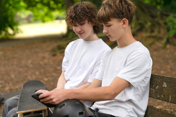 Two boys enjoy downtime at a skate park, discussing tricks and checking equipment while resting on a bench in summer