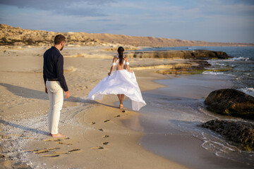 Bride in flowing white dress walks towards the sea as groom watches, a tender moment captured on a rocky coastline.