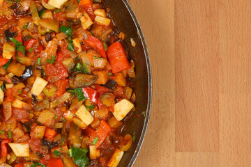 A top-down close-up view of a rustic Italian caponata, a delicious stew of eggplant and other vegetables in a pan, with a wooden background and copy space