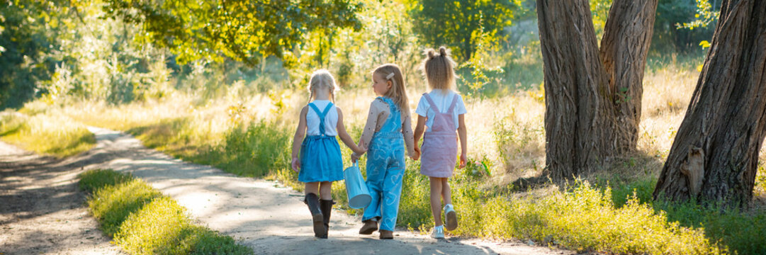 Three little girls holding hands walking with watering can outdoors. Concept of eco-education, childhood friendship, family care and sustainable life. Banner