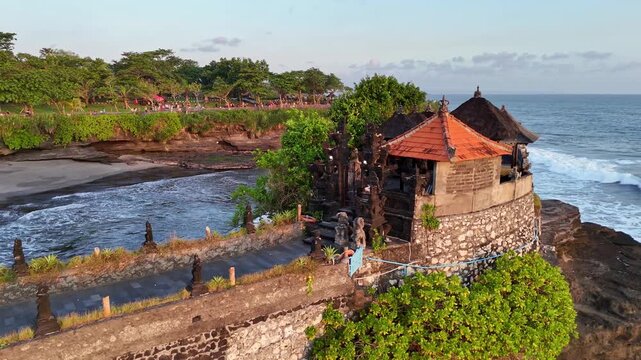 Drone view of Batu Bolong Temple perched on a rocky formation extending into the ocean, surrounded by the ocean, small temples, and people walking along the paved area. Location: Bali, Indonesia