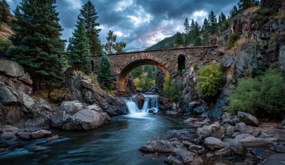 A rustic stone arch bridge spans a rocky mountain stream, framed by towering pines and dramatic clouds
