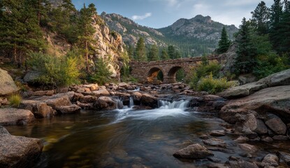 Rocky mountain stream flows under an arched stone bridge. Sunlight bathes the scene