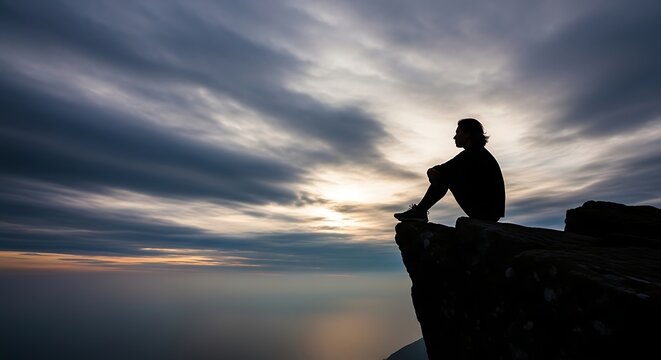 A solitary figure in silhouette sits on a rocky cliff, looking out over the sea towards a dramatic, cloudy sunset. - Powered by Adobe