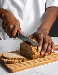 A loaf of whole grain bread is sliced on a light wooden cutting board by a pair of hands.