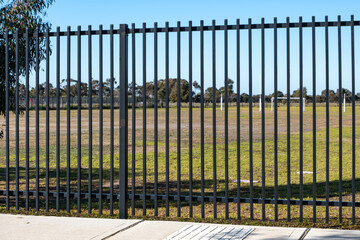 Close-up view of a tall black vertical metal fence enclosing an open grassy sports field in...
