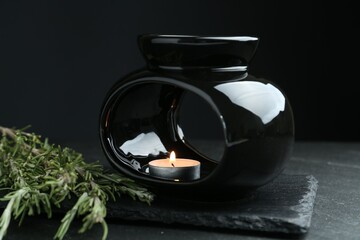 Aroma lamp with burning candle and rosemary on dark textured table against black background, closeup