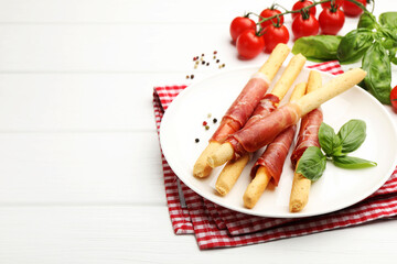 Delicious grissini sticks with prosciutto, peppercorns and basil on white wooden table, closeup. Space for text